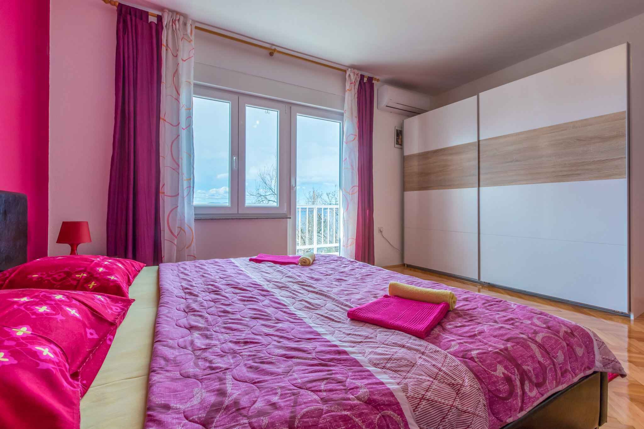 Une chambre avec un grand lit, une literie rose, une fenêtre et une armoire.
