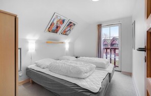 Quarto com cama, mesa de cabeceira, luminária na parede e janela com vista para um edifício.