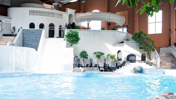Ein Innenpool mit Wasserfall, Sitzbereichen und einer Treppe, die in eine erhöhte Ebene führt.