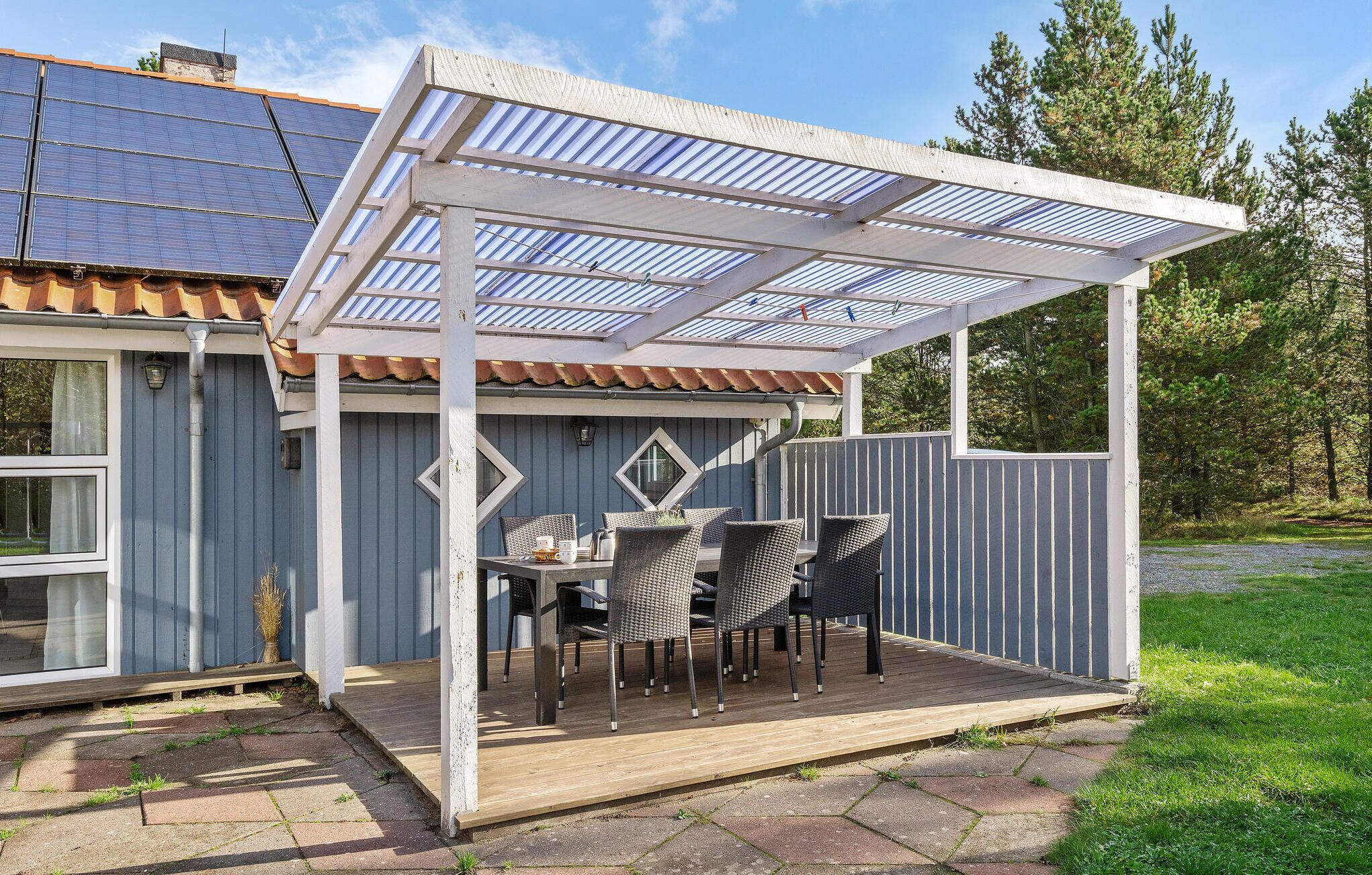 A covered patio with outdoor furniture, a pergola with solar panels, and a view of a house with a blue exterior.