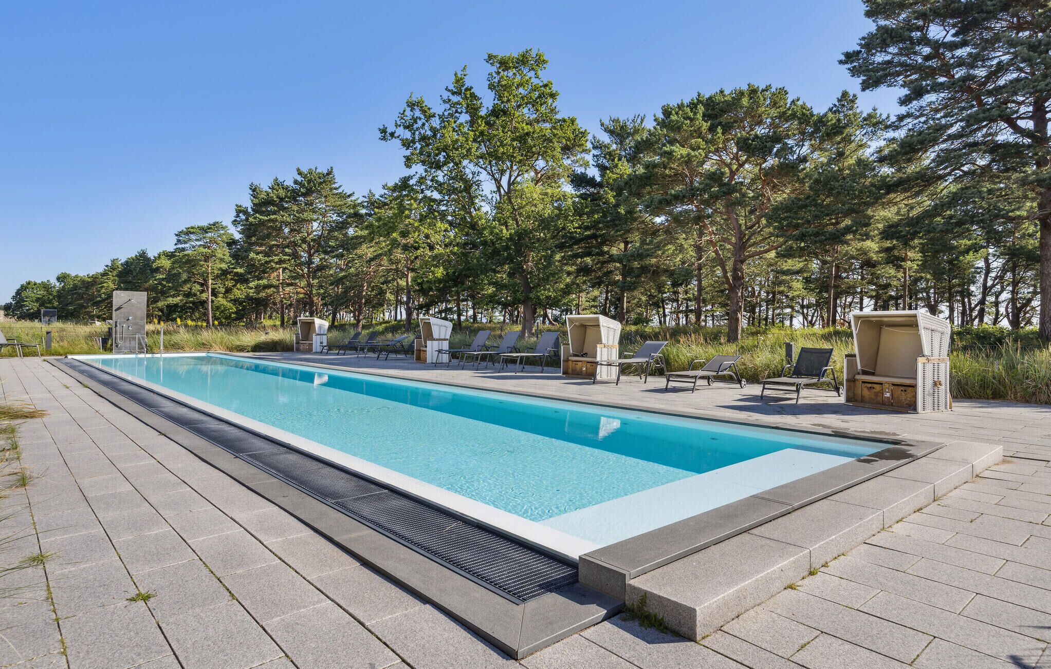 Ein rechteckiger Pool mit klarer, blauer Wasseroberfläche, umgeben von einer gepflasterten Liegefläche. Mehrere Liegestühle und Sonnenliegen sind um den Pool herum aufgestellt, und es befindet sich eine kleine Holzkonstruktion mit einer Bank in der Nähe.