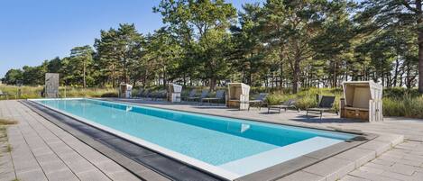 Ein rechteckiger Pool mit klarer, blauer Wasseroberfläche, umgeben von einer gepflasterten Liegefläche. Mehrere Liegestühle und Sonnenliegen sind um den Pool herum aufgestellt, und es befindet sich eine kleine Holzkonstruktion mit einer Bank in der Nähe.