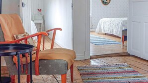 Une pièce chaleureuse avec une chaise en bois, une petite table, un tapis rayé aux couleurs vives et un lit recouvert d’une courtepointe blanche.