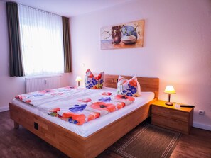 Quarto com cama de madeira, mesa de cabeceira com luminária, radiador e janela com cortinas.