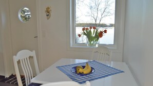 Et spiseområde med et bord dækket til to personer, et vindue med udsigt over en sø, og en vase med blomster på vindueskarmen.
