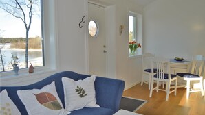 Et værelse med blå sofa, hvide puder, et spisebord med stole, et vindue med søudsigt og en dør med knagerække.