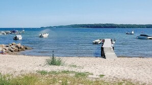 Una playa con un muelle de madera que se adentra en aguas tranquilas, rodeada de barcos y una orilla con vegetación.