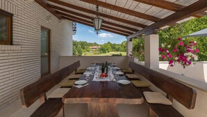 Un espace extérieur aménagé pour les repas, avec une longue table en bois pouvant accueillir six personnes, des bancs de chaque côté, et une vue sur la verdure et une maison au loin.