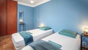 A room with two beds, each with a blue blanket and patterned pillow, a small table with a lamp, a mirror, and a wooden wardrobe. - 3 bedroom nice home in Saint-Thomé (Saint-Thomé)