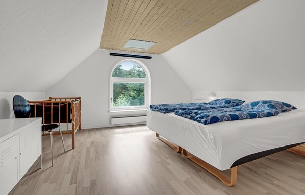 A bedroom with a sloped ceiling, a large window, a bed with blue patterned bedding, a desk with a chair, and a wardrobe.