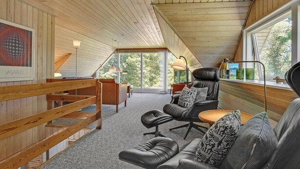 Ein gemütliches Loft mit schrägverdeckter Decke, Holzwänden und großen Fenstern mit Blick auf Grünflächen.