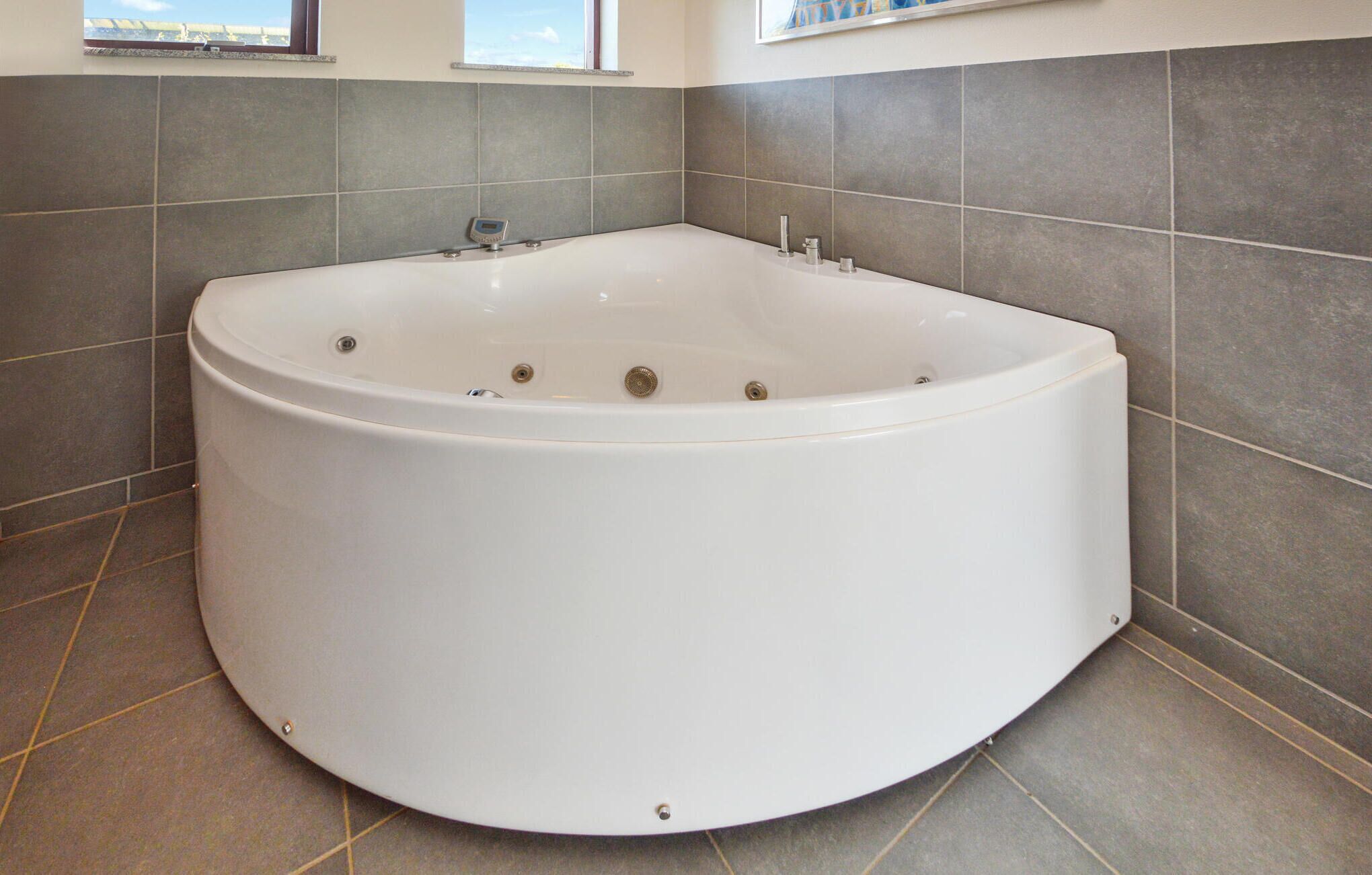 A large, round, white bathtub in a bathroom with grey tiles and a window above it.