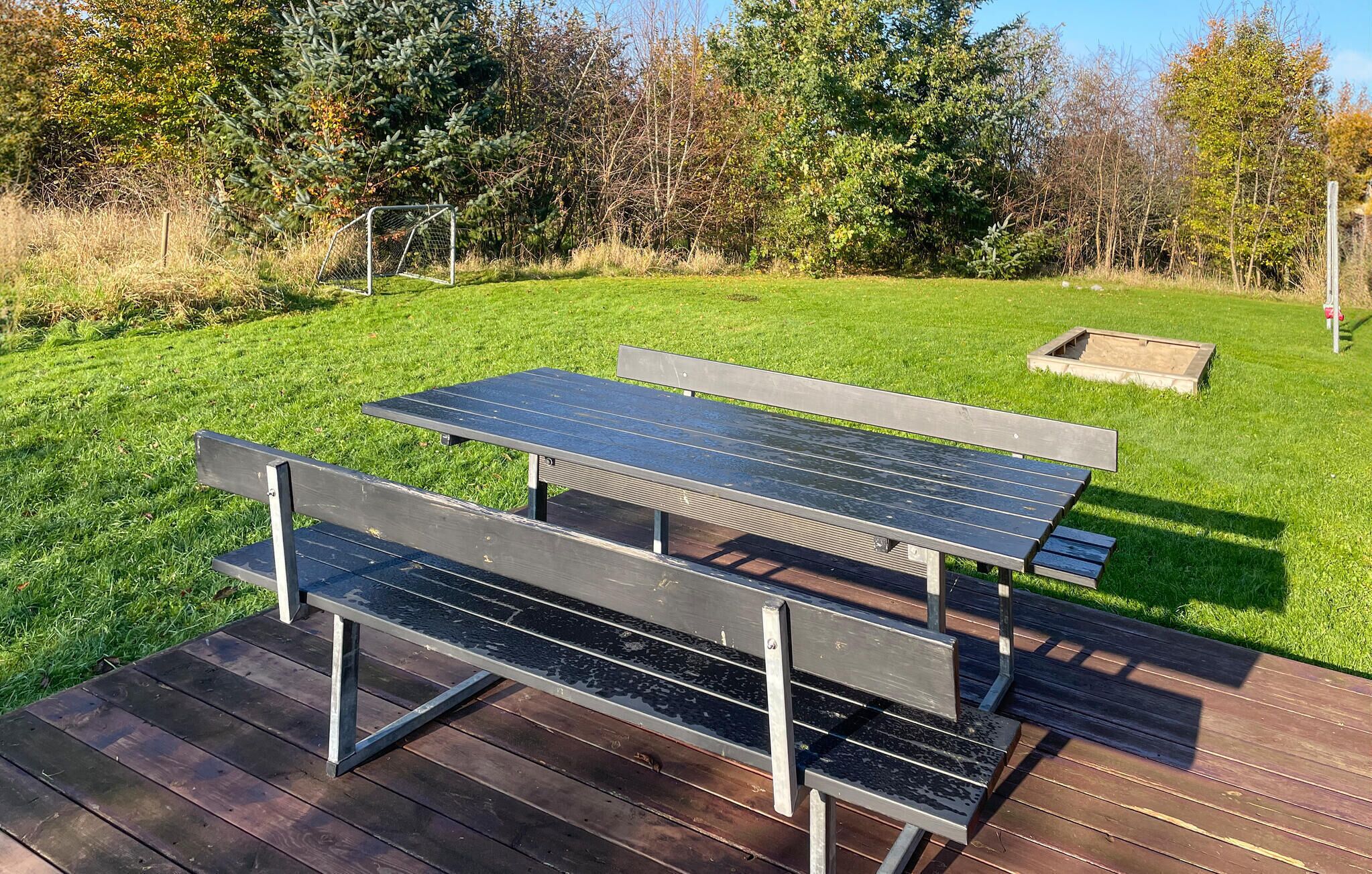 A picnic table with benches on a wooden deck, surrounded by a grassy area with trees and a fence in the background.