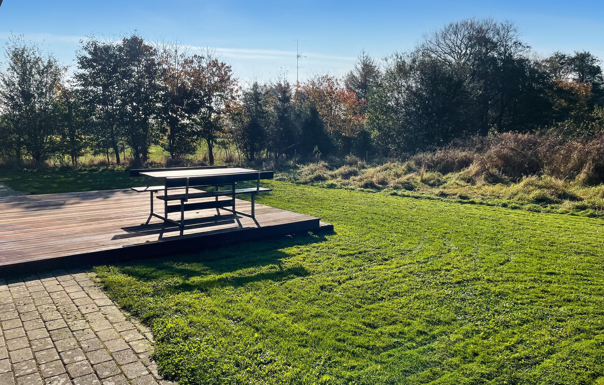 Et picnicbord på en træterrasse med græs og træer i baggrunden.