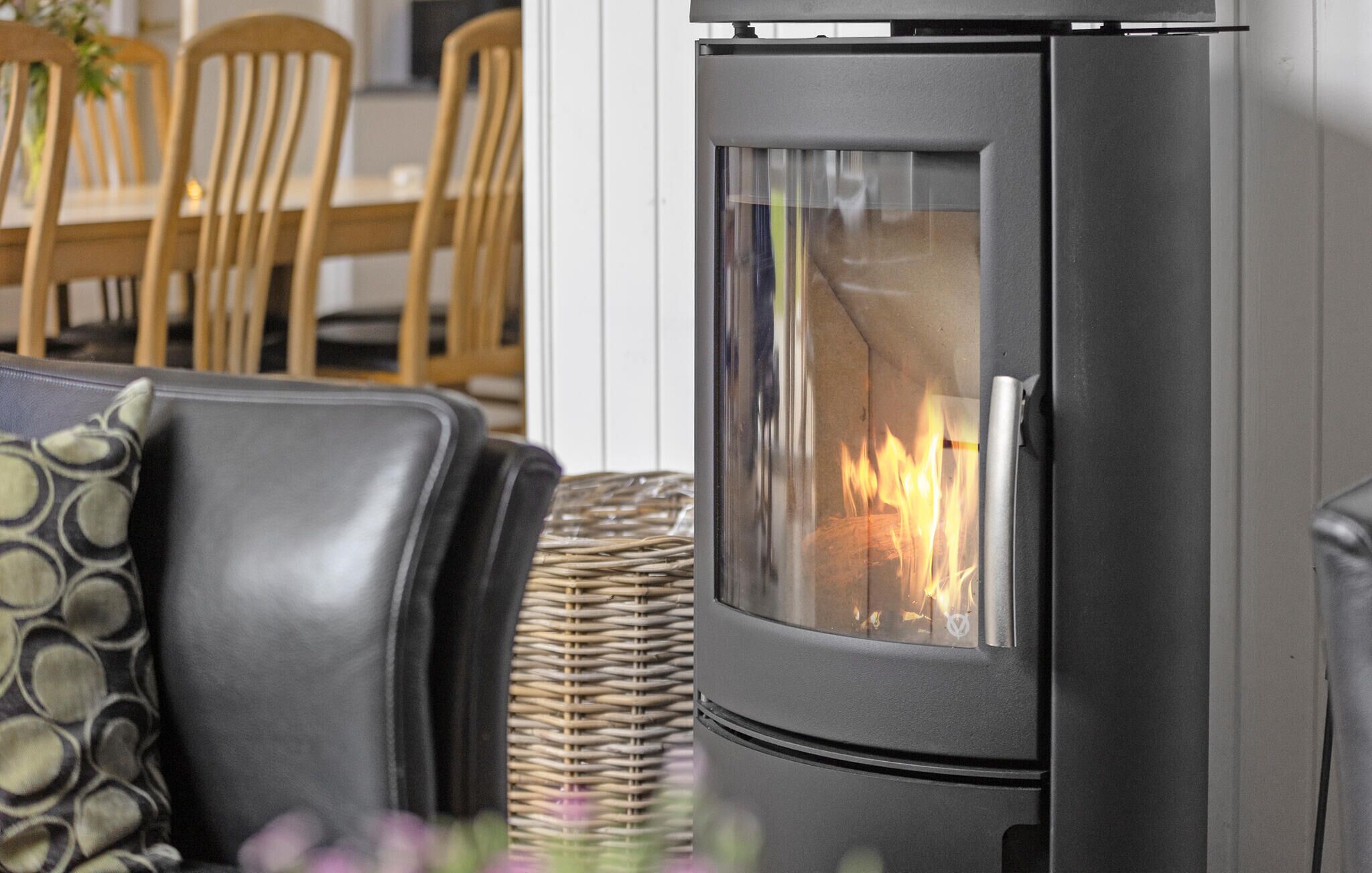 A modern fireplace with a glass door, a wicker basket, and a dining area with wooden chairs and a table.