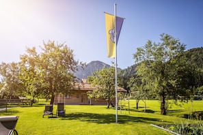 A flagpole with a flag, a building with a red-tiled roof, and a grassy area with trees and a mountain in the background. - Double room Primerl - Anderlbauer am See (Schliersee)