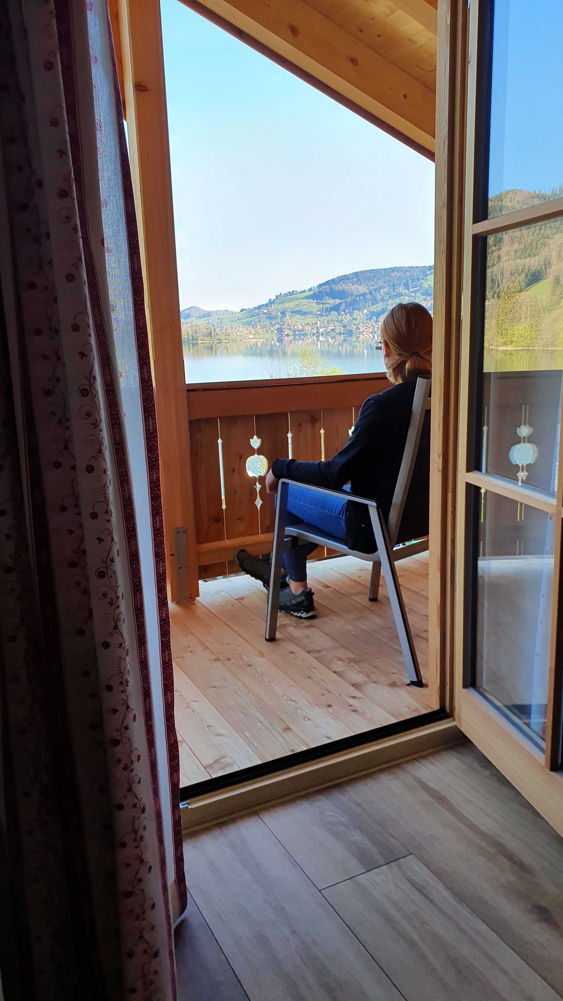 A person seated on a chair on a wooden deck with a view of a lake and hills.