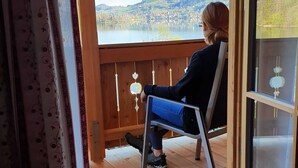 A person seated on a chair on a wooden deck with a view of a lake and hills. - Double room Primerl - Anderlbauer am See (Schliersee)