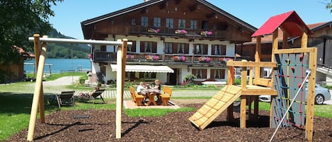 A playground with swings and climbing structures, a table with chairs, and a building with balconies and flower boxes in the background.