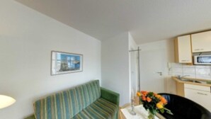 A small, compact living space with a sofa, a dining table with a vase of flowers, and a kitchenette in the background. - Villa Aquamarina Whg. 18, SOUTH FACING BALCONY, CHAIR - Villa Aquamarina, 1st ROW, partly. (Heringsdorf)
