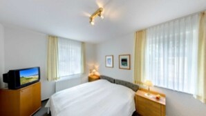 A hotel room with a bed, a TV on a wooden cabinet, a bedside table with a lamp, and two framed pictures on the wall. - Villa Aquamarina Whg. 18, SOUTH FACING BALCONY, CHAIR - Villa Aquamarina, 1st ROW, partly. (Heringsdorf)