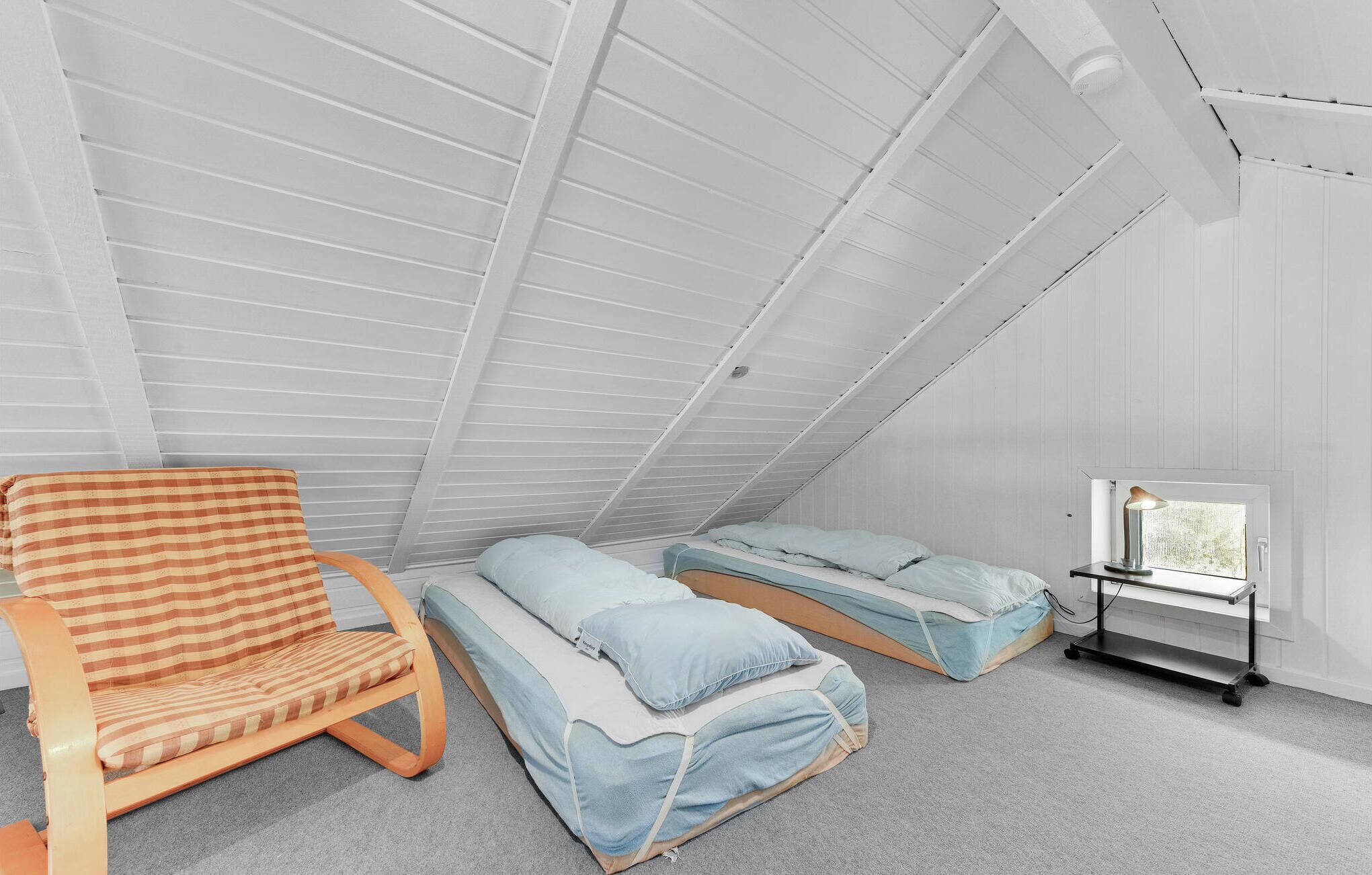 A room with a sloped ceiling, a wooden chair, two beds with light blue bedding, and a table lamp.