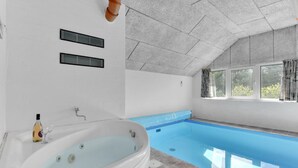 A modern bathroom with a large bathtub, a window with curtains, and a pool area. - Lovely home in Ringkøbing with sauna (Ringkøbing)