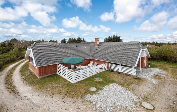 A two-story brick house with a sloped roof, a fenced patio area with outdoor furniture, and a gravel driveway.