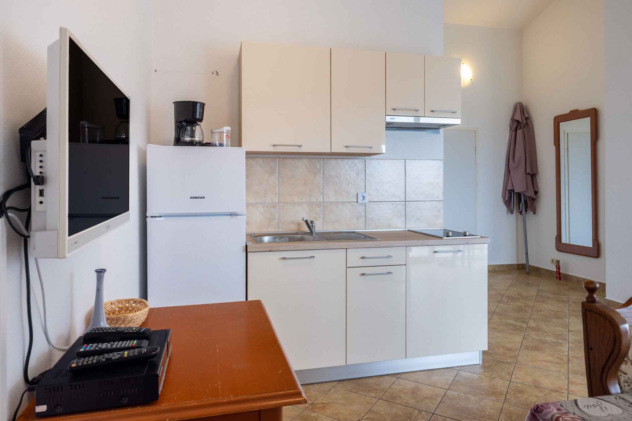 Cocina compacta con armarios blancos, horno empotrado y lavamanos. Hay una mesa de madera con una cesta y una cafetera encima.