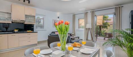 A modern dining area with a table set for breakfast, a kitchen in the background, and a view of the outdoors.