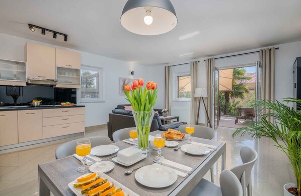 A modern dining area with a table set for breakfast, a kitchen in the background, and a view of the outdoors.