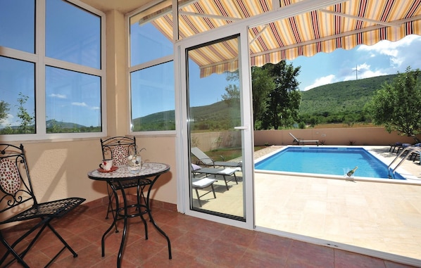 A sunlit patio with a pool, a table with chairs, and a view of a hilly landscape.