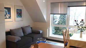 Una sala moderna con un sofá gris oscuro, una mesa de centro de madera y una mesa de comedor con sillas. Hay dos obras de arte enmarcadas en la pared, una ventana con persianas y un jarrón con flores sobre la mesa de comedor.