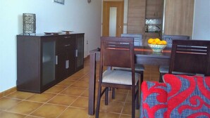 A dining area with a dark wooden cabinet, two chairs, a table with a bowl of fruit, and a tiled floor.