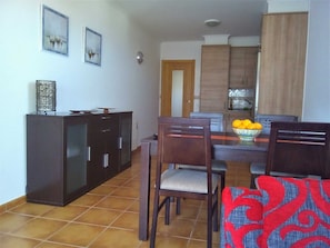 A dining area with a dark wooden cabinet, two chairs, a table with a bowl of fruit, and a tiled floor.