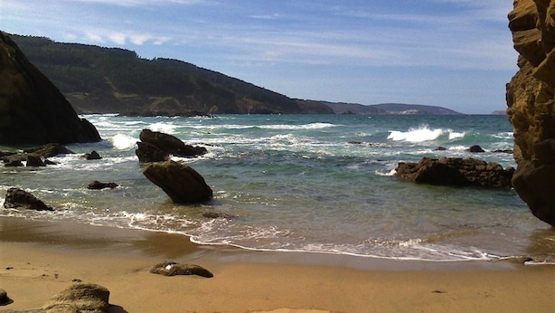 A beach with clear water, rocks, and a hilly landscape in the background.