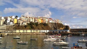 A coastal town with a harbor filled with boats, a prominent hill with colorful buildings, and a stone wall along the waterfront.