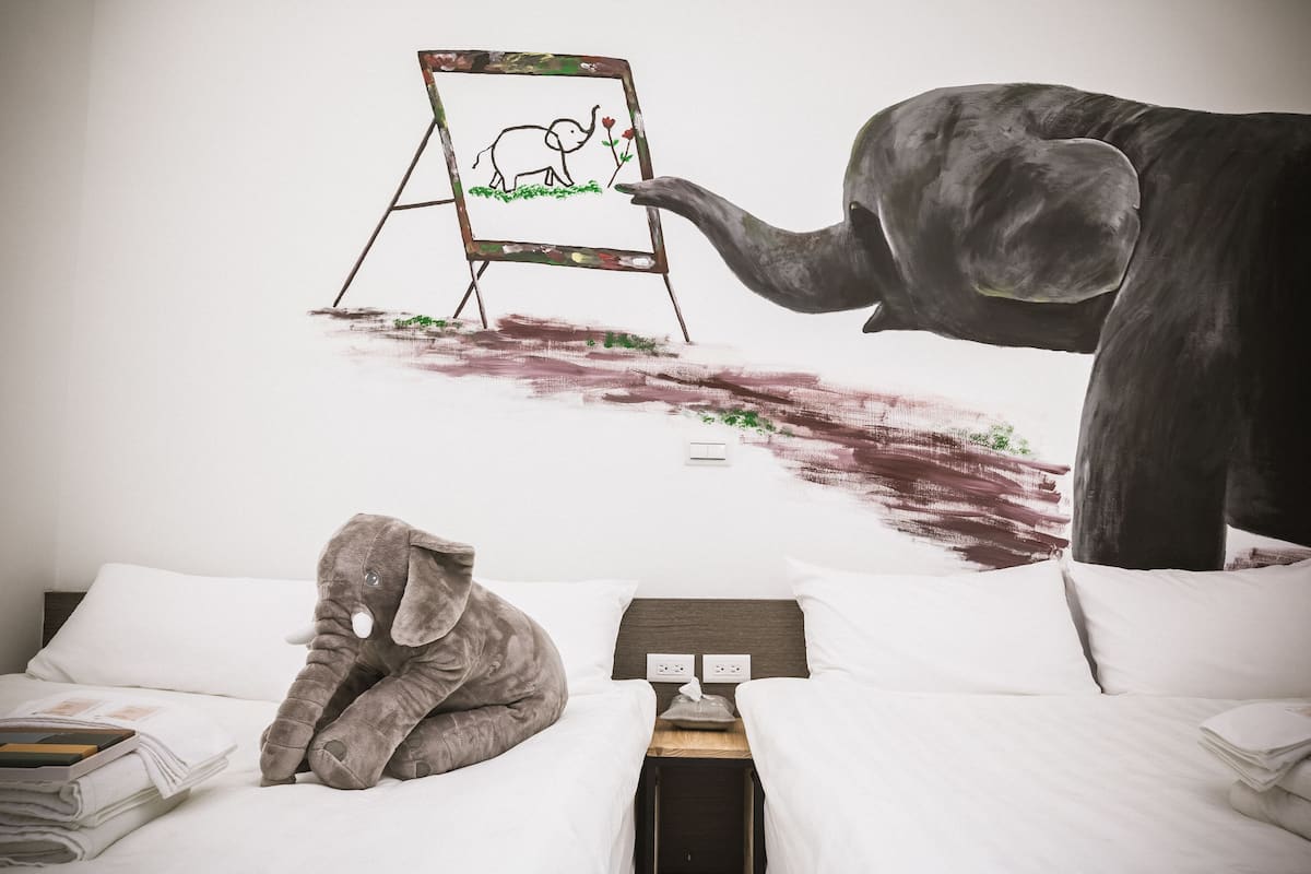 A bedroom with a bed, pillows, a nightstand, and a wall mural of an elephant painting on an easel.