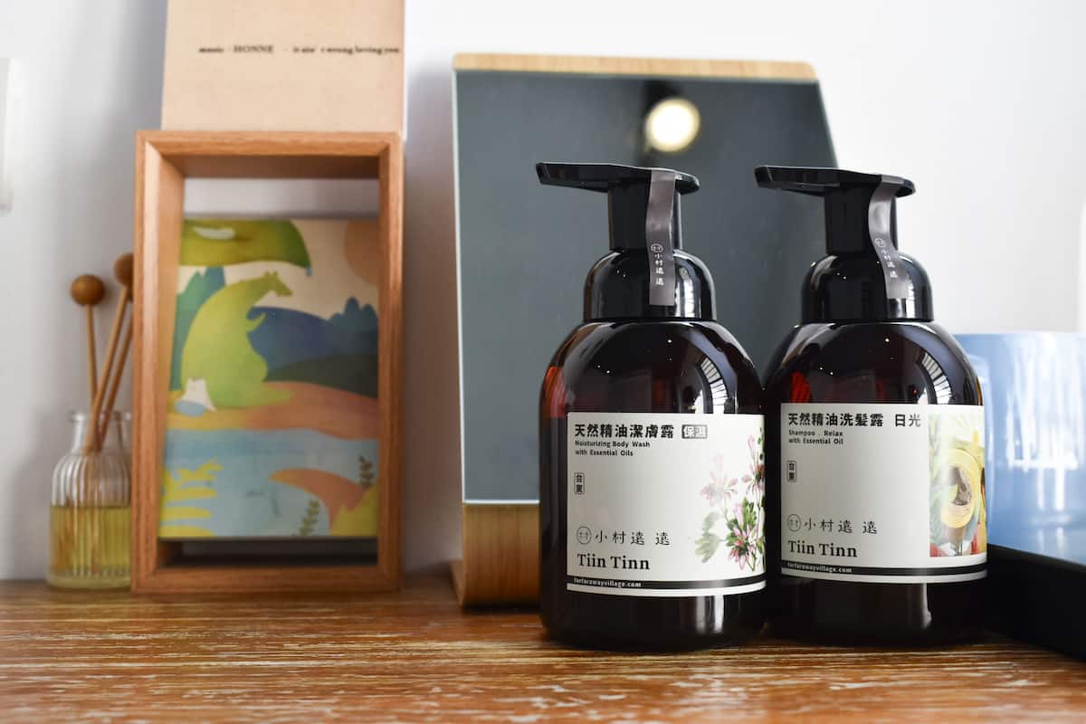 Two bottles of Tinn body wash and shampoo on a wooden surface, with a framed picture and a small bottle of oil in the background.