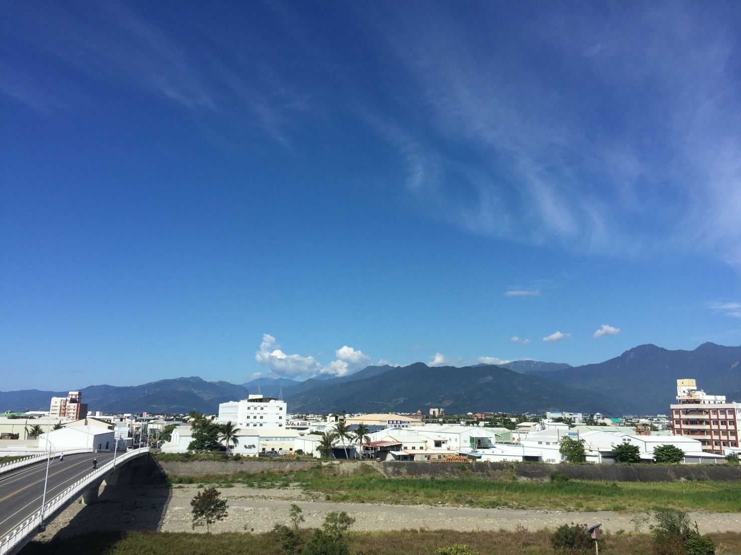 A bridge with a road passing over a green area, leading to a town with white buildings and mountains in the background.