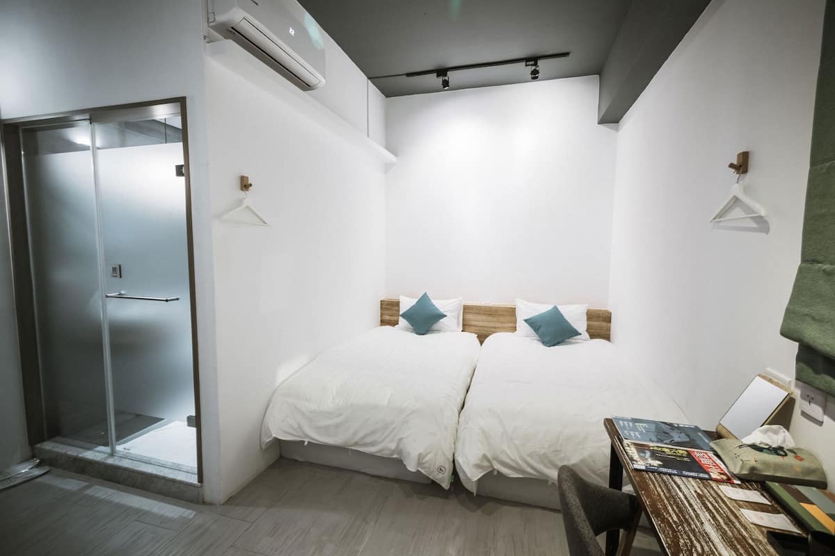 A room with a bed, a desk with items, a shower, and an air conditioning unit.
