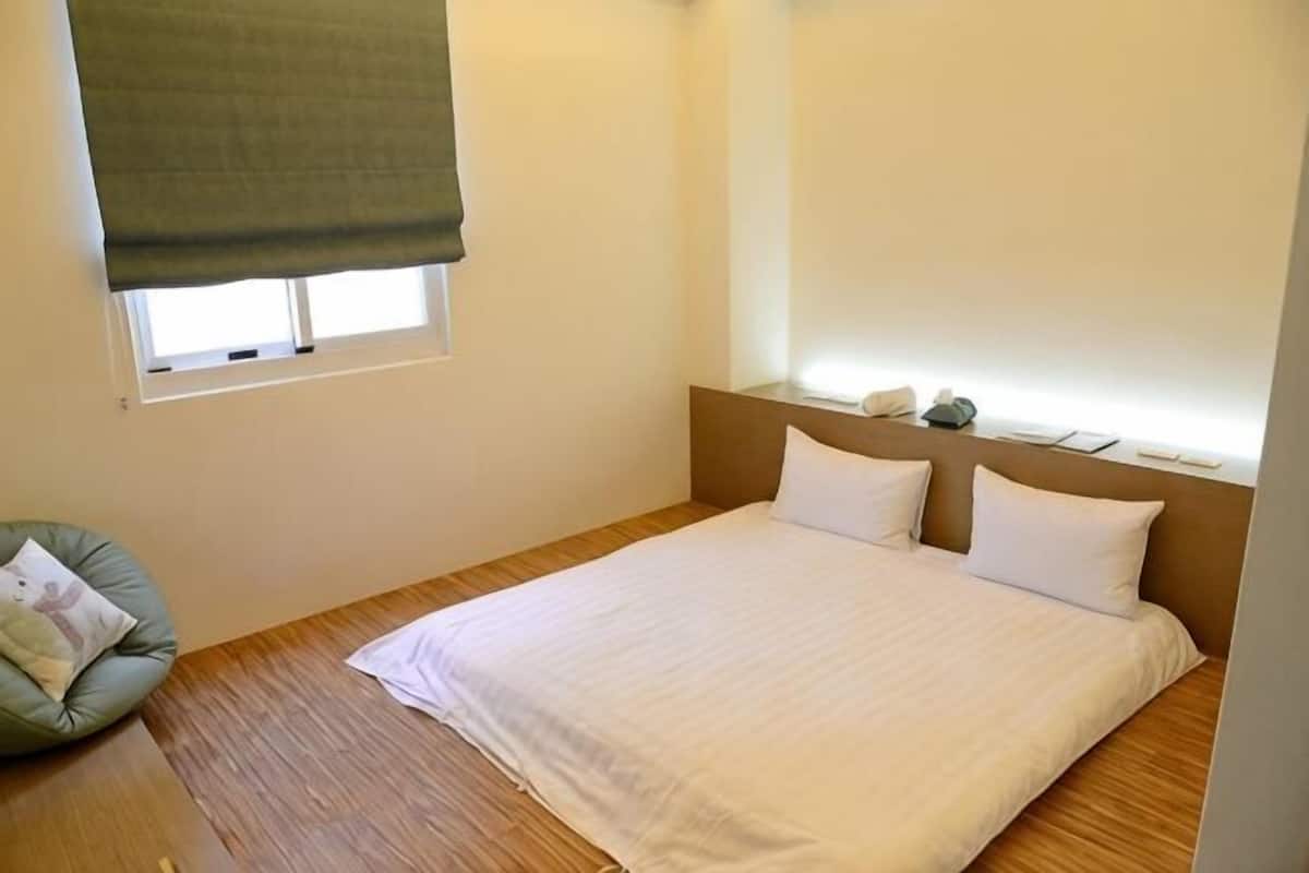 A bedroom with a bed, a window with a blind, a bedside table, and a chair.