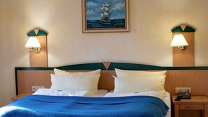 Een net opgemaakt bed met een blauwe deken, twee bedlampjes en een schilderij van een schip aan de muur.