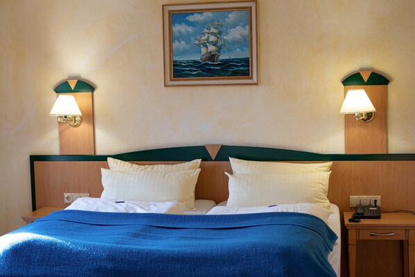 Una cama bien hecha con un edredón azul, dos lámparas de noche y un cuadro de un barco en la pared.