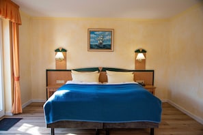 Une chambre d’hôtel avec un grand lit, deux tables de chevet, un tableau représentant un navire accroché au mur et une fenêtre avec des rideaux.