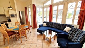 A living room with a sofa, chairs, a glass coffee table, and a dining table with chairs. There are windows with curtains, a kitchen area with a microwave, and a refrigerator.