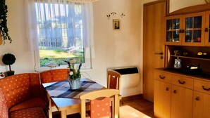 Un coin repas chaleureux avec une table dressée pour deux personnes, un buffet en bois aux portes vitrées et une fenêtre donnant sur l’extérieur.