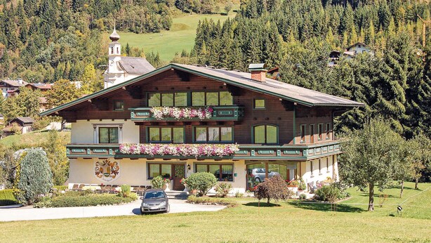 En toetages chalet med balkon, omgivet af en velplejet have og et bjergrigt landskab.