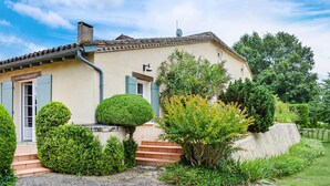 Uma casa de dois andares com fachada bege, persianas verdes e um pequeno jardim com arbustos variados e uma árvore.