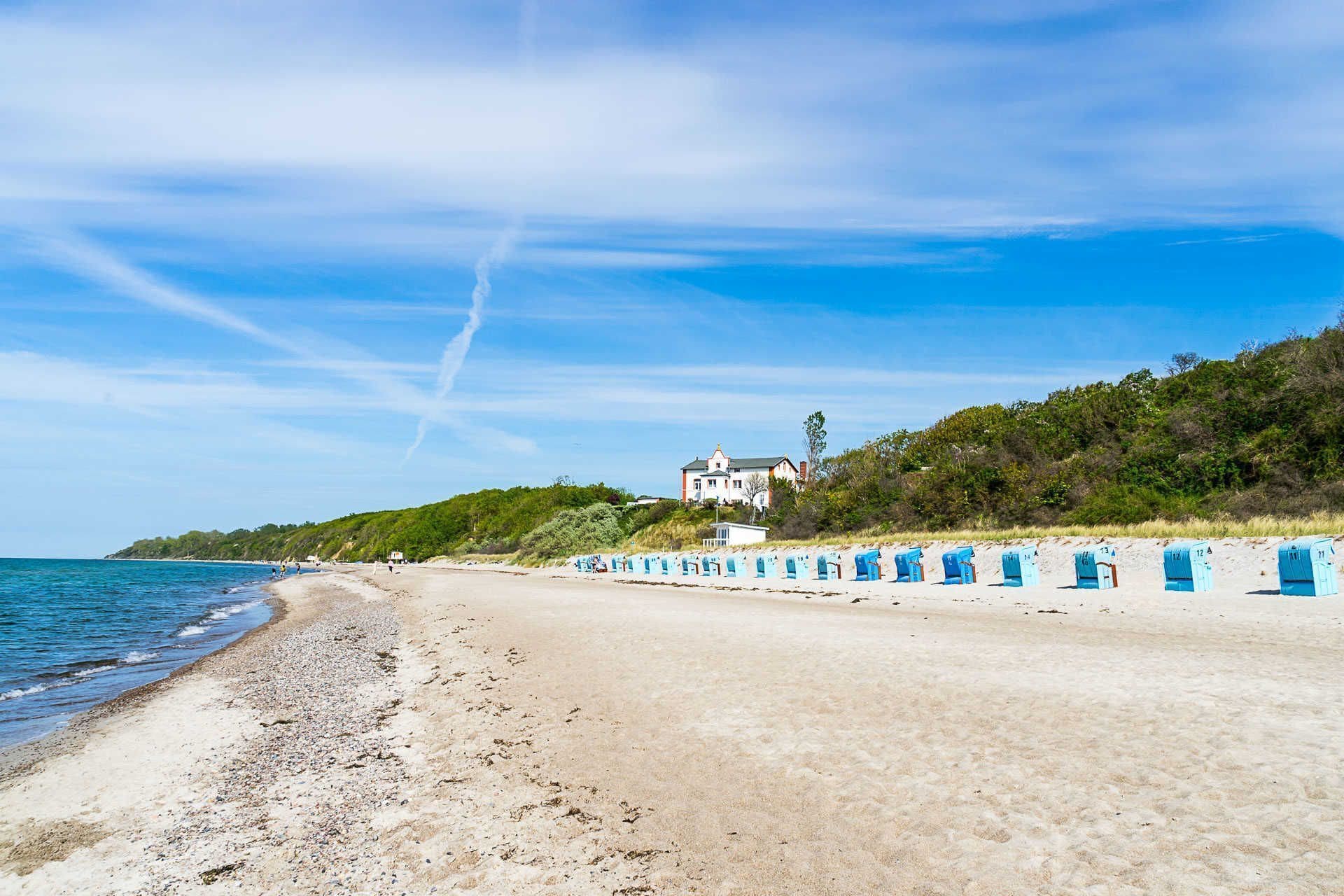 Une plage bordée d’une rangée de cabanes de plage bleues, une grande maison située sur une colline et un ciel dégagé.